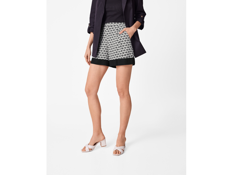 Woman in dark jacket, patterned shorts, and light-colored low-heeled mules.