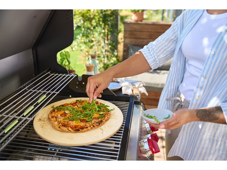 Woman topping a grilled pizza with arugula on a pizza stone outdoors.