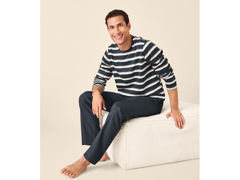 Smiling man in striped pyjamas and dark blue trousers, sitting on a white pouffe.