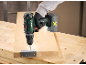 Gloved worker using a cordless drill to attach a hinge to a wooden board.