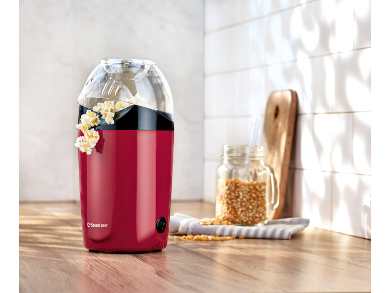 Red popcorn maker with popcorn on a wooden countertop, next to a jar of corn kernels.