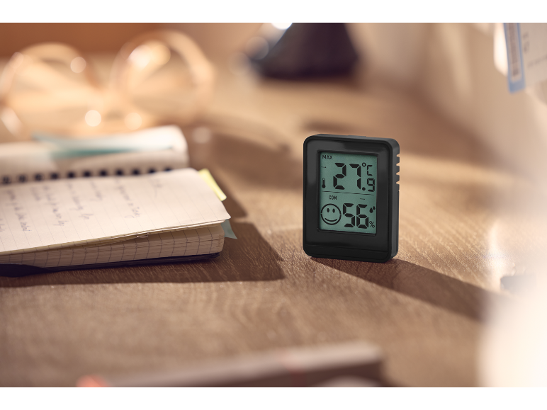 Digital thermo-hygrometer on a wooden desk next to an open notebook.