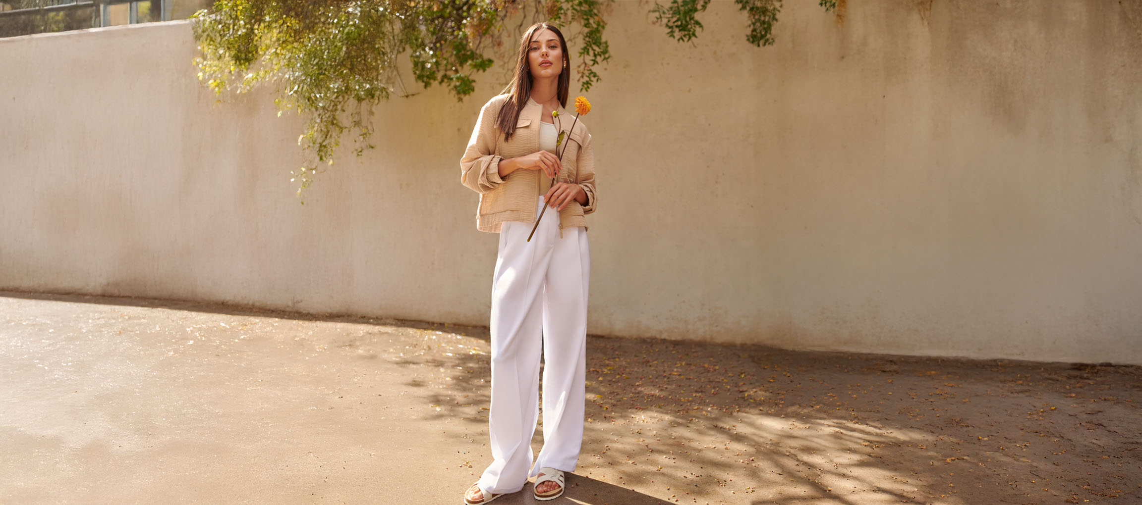 Woman in a beige jacket, white trousers, and sandals, holding a yellow flower.