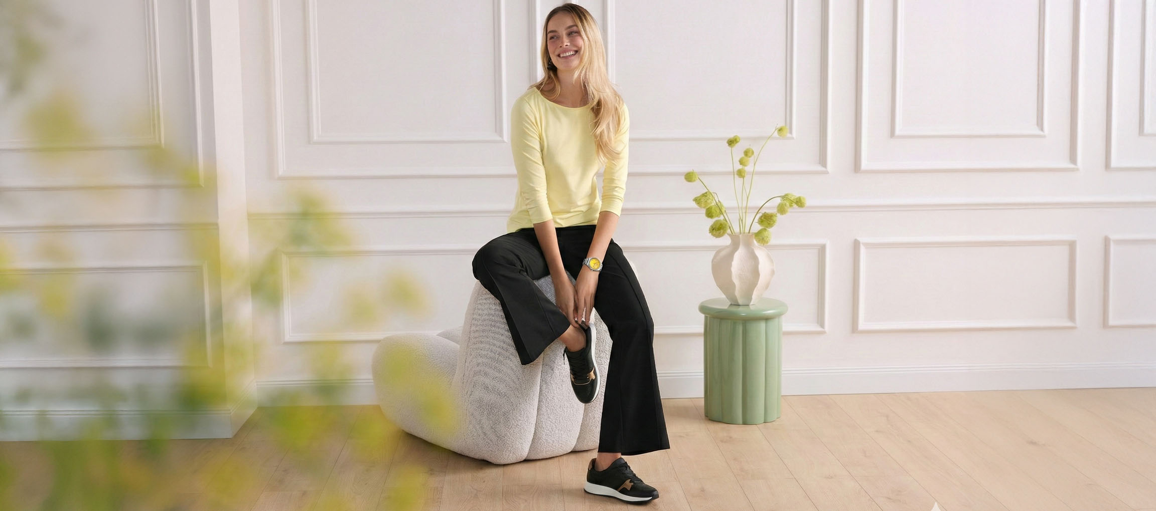 Woman in yellow top and black trousers sitting on a pouf, smiling.