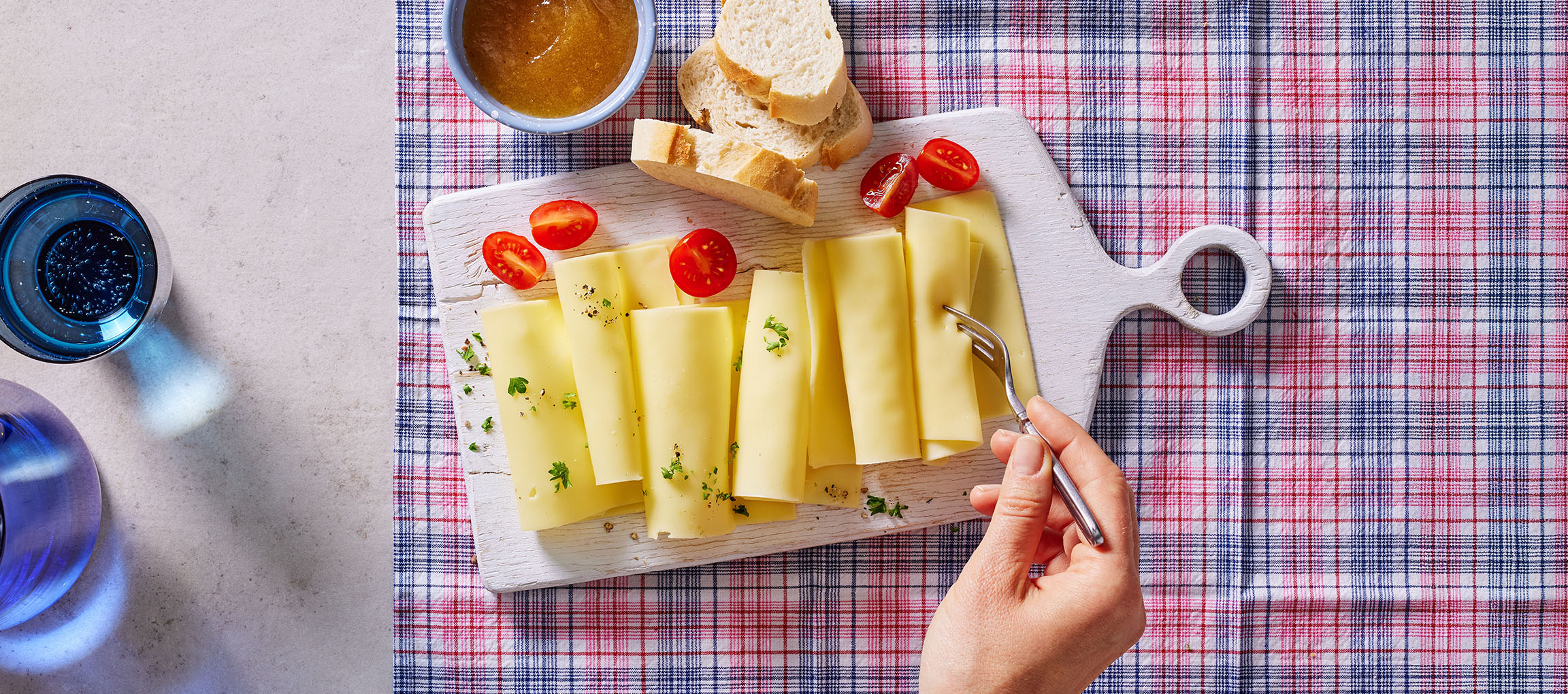 Cheese slices with cherry tomatoes, bread, and jam on a checkered tablecloth.