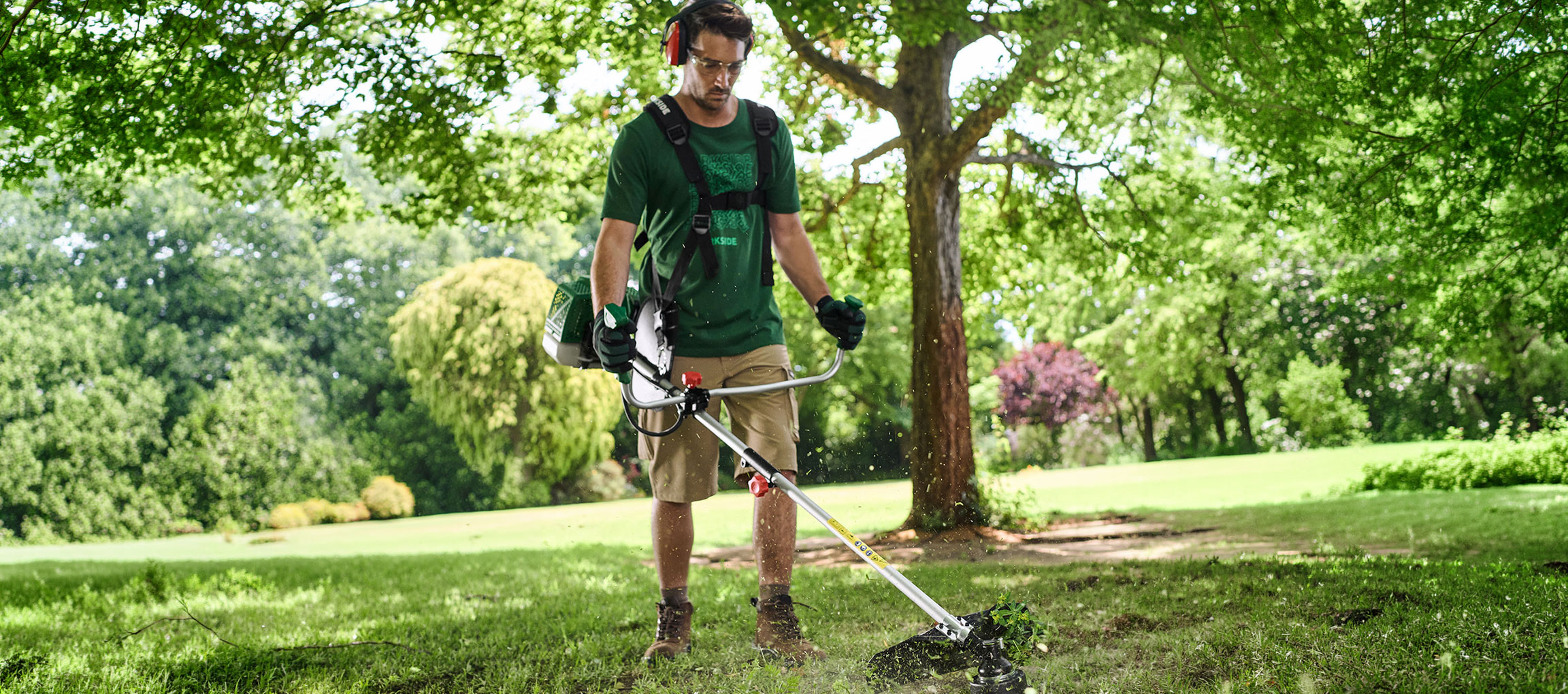 Man with a brush cutter and protective gear trimming grass in a garden.