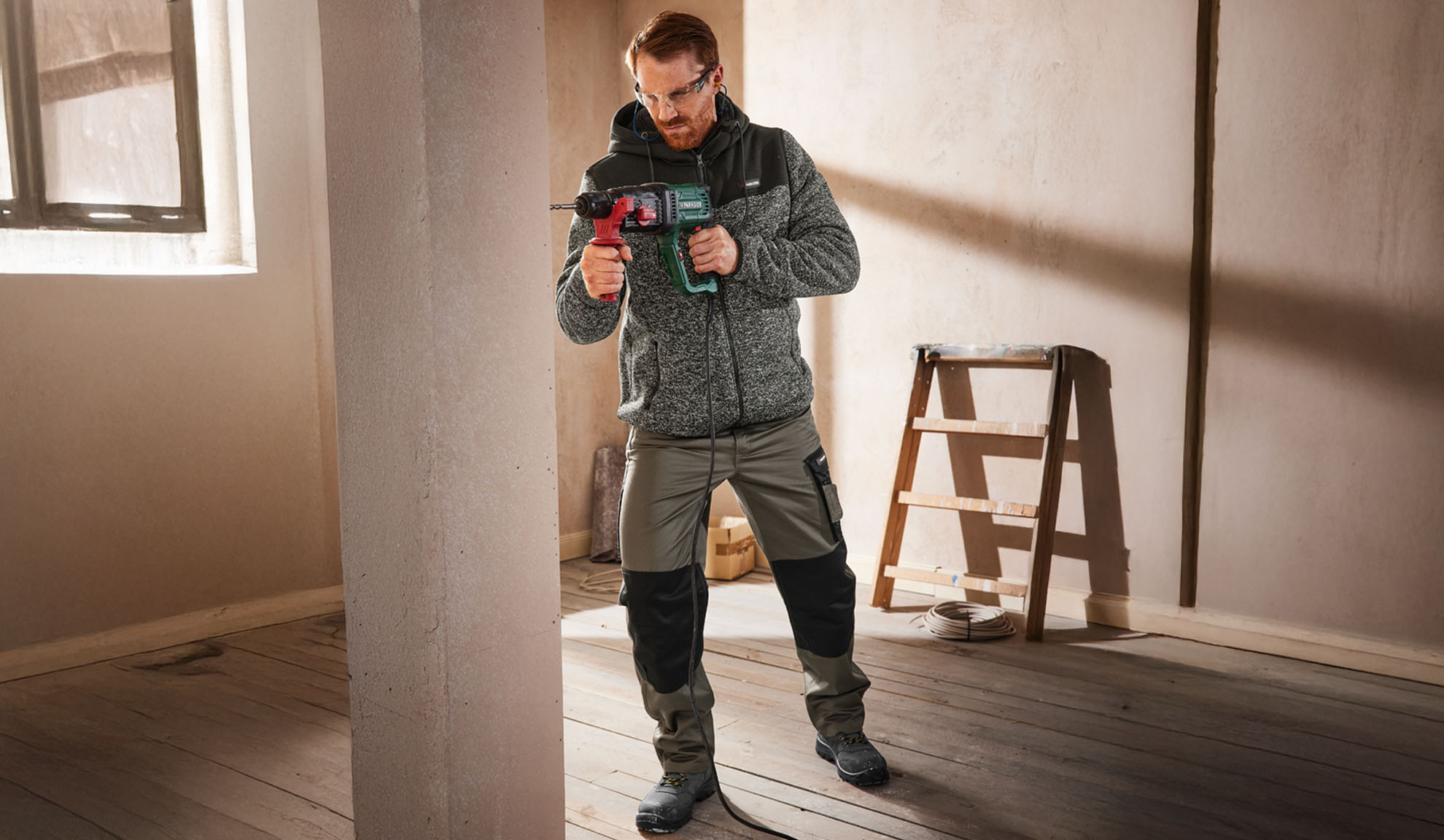 Man in work jacket and trousers, holding a drill in an unfinished room.