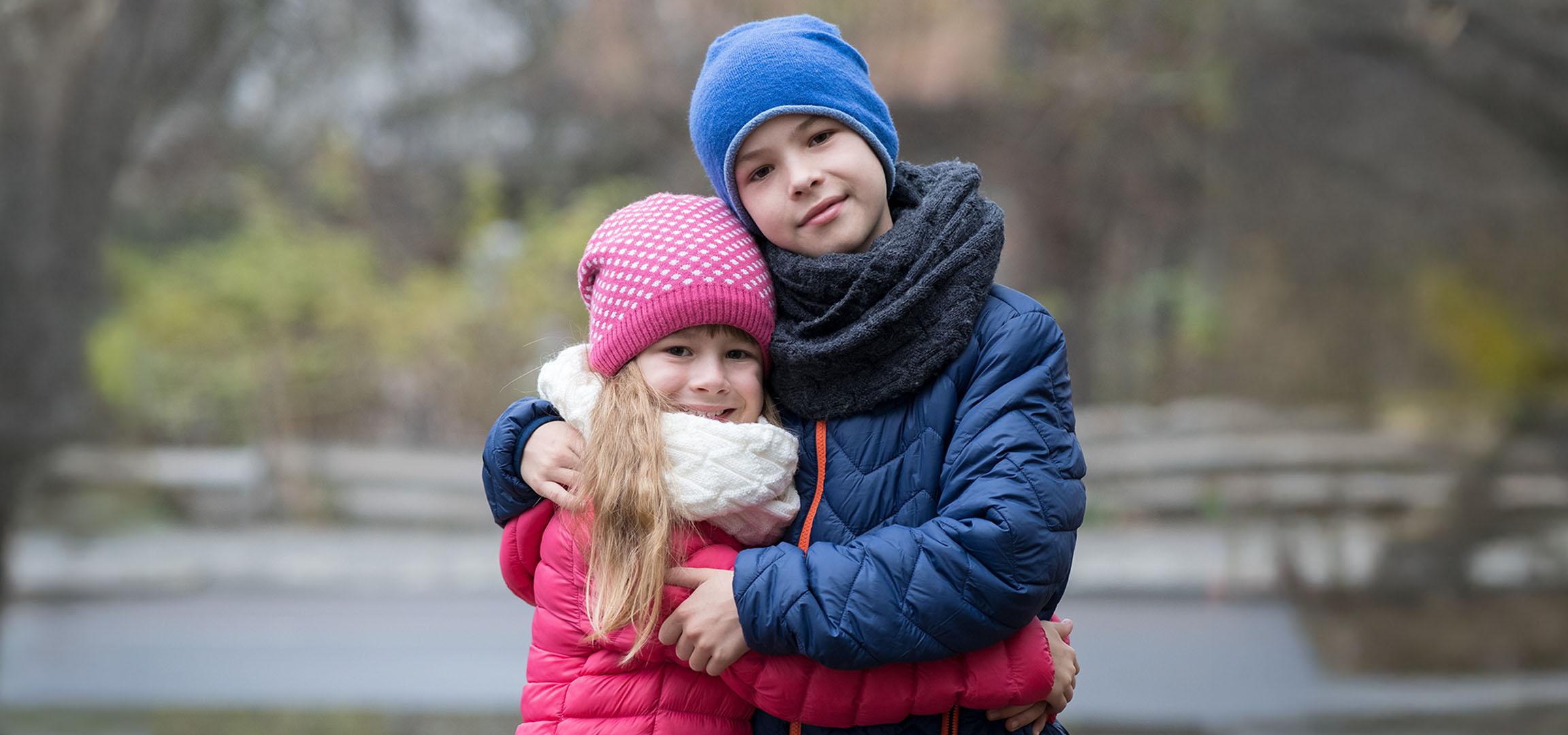 Two children in winter hats and jackets hugging outdoors.