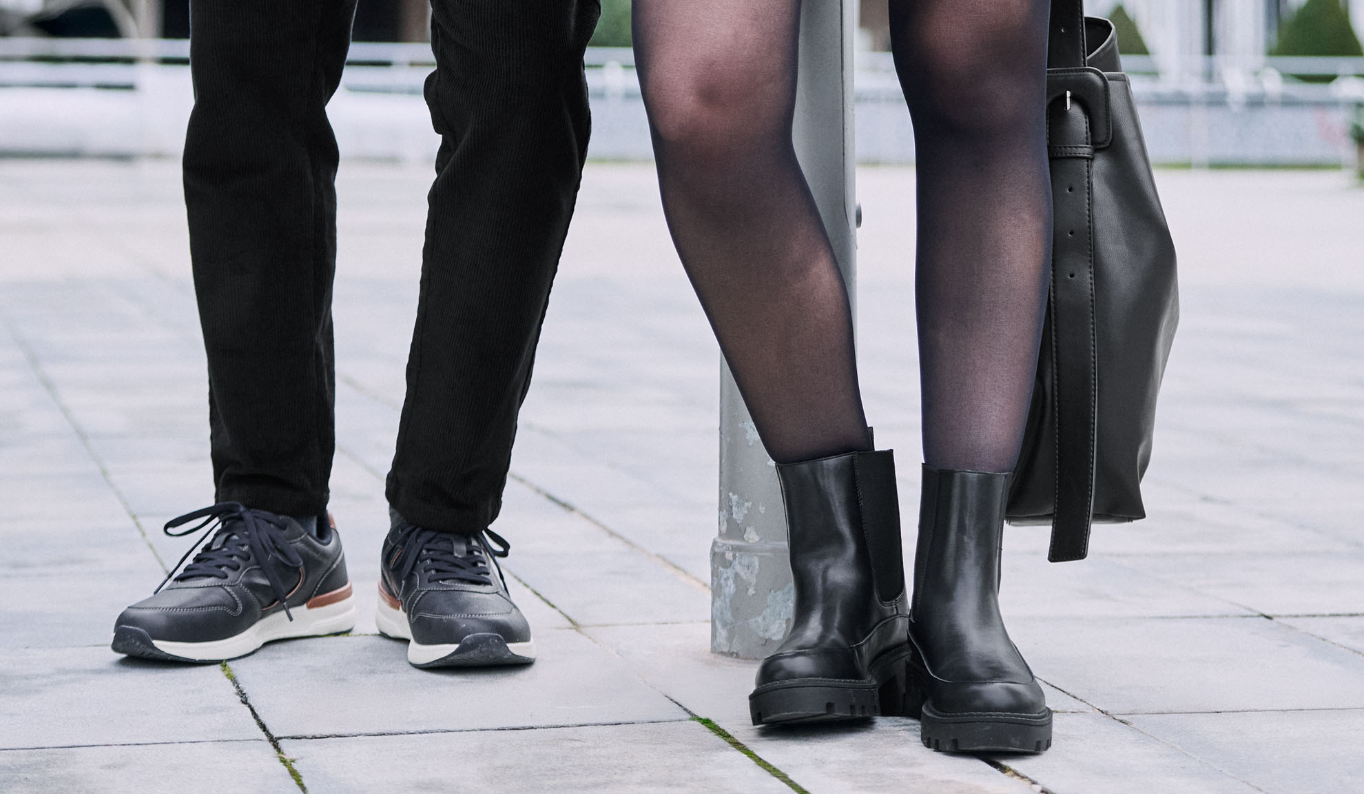 Legs of two people, one wearing pants and sneakers, the other tights, boots, and a bag.