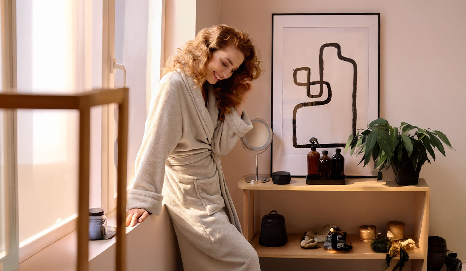 A woman in a bathrobe by a window, with a shelf of beauty products and a plant.