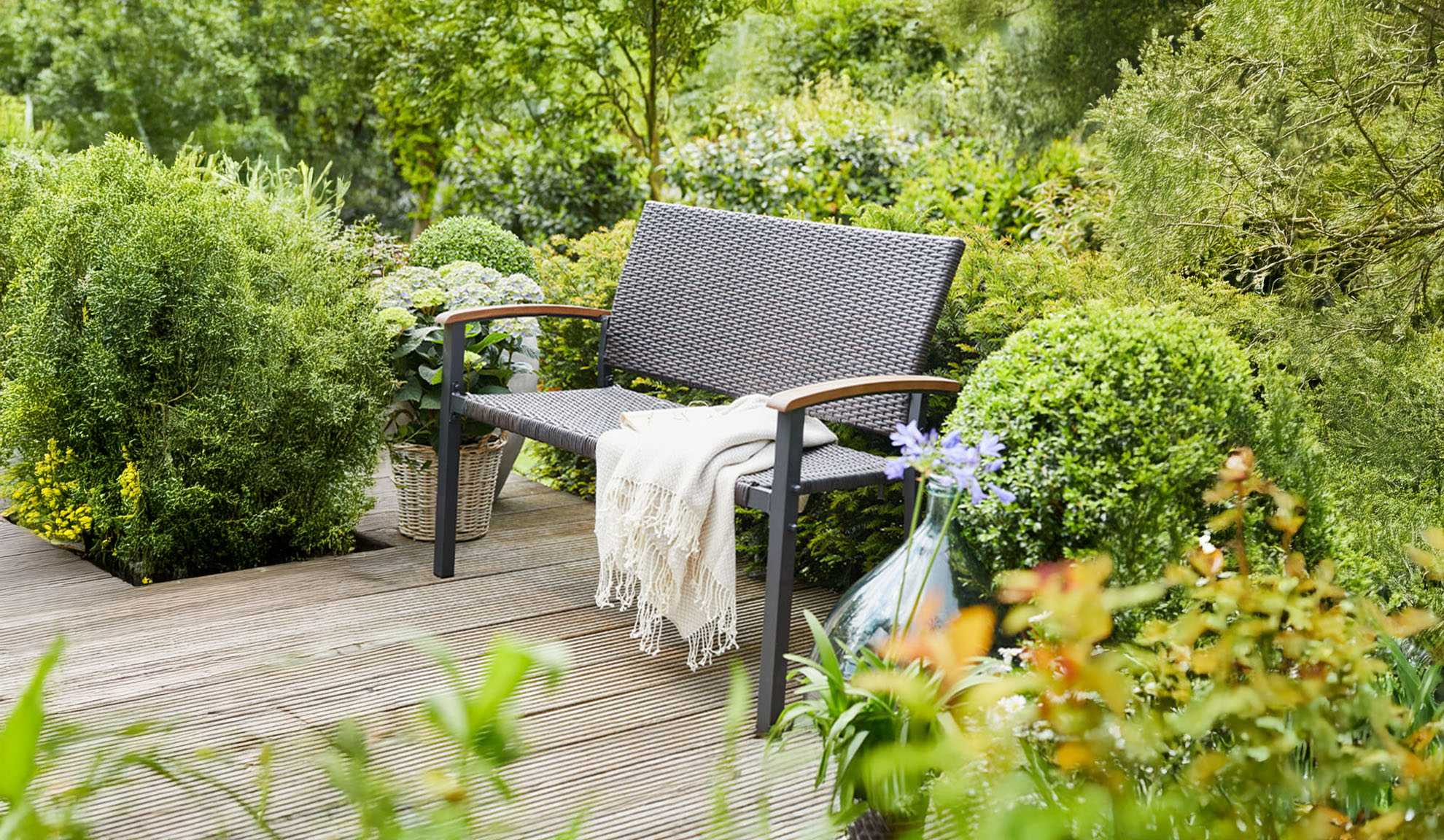 Rattan garden bench on a wooden deck, surrounded by lush greenery.