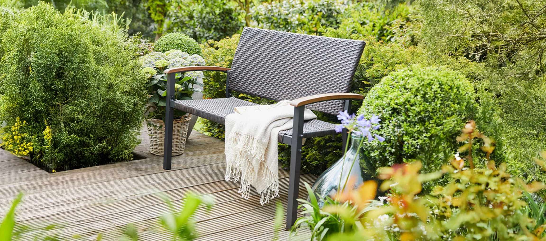 A gray garden bench with a blanket on a wooden deck surrounded by green plants.