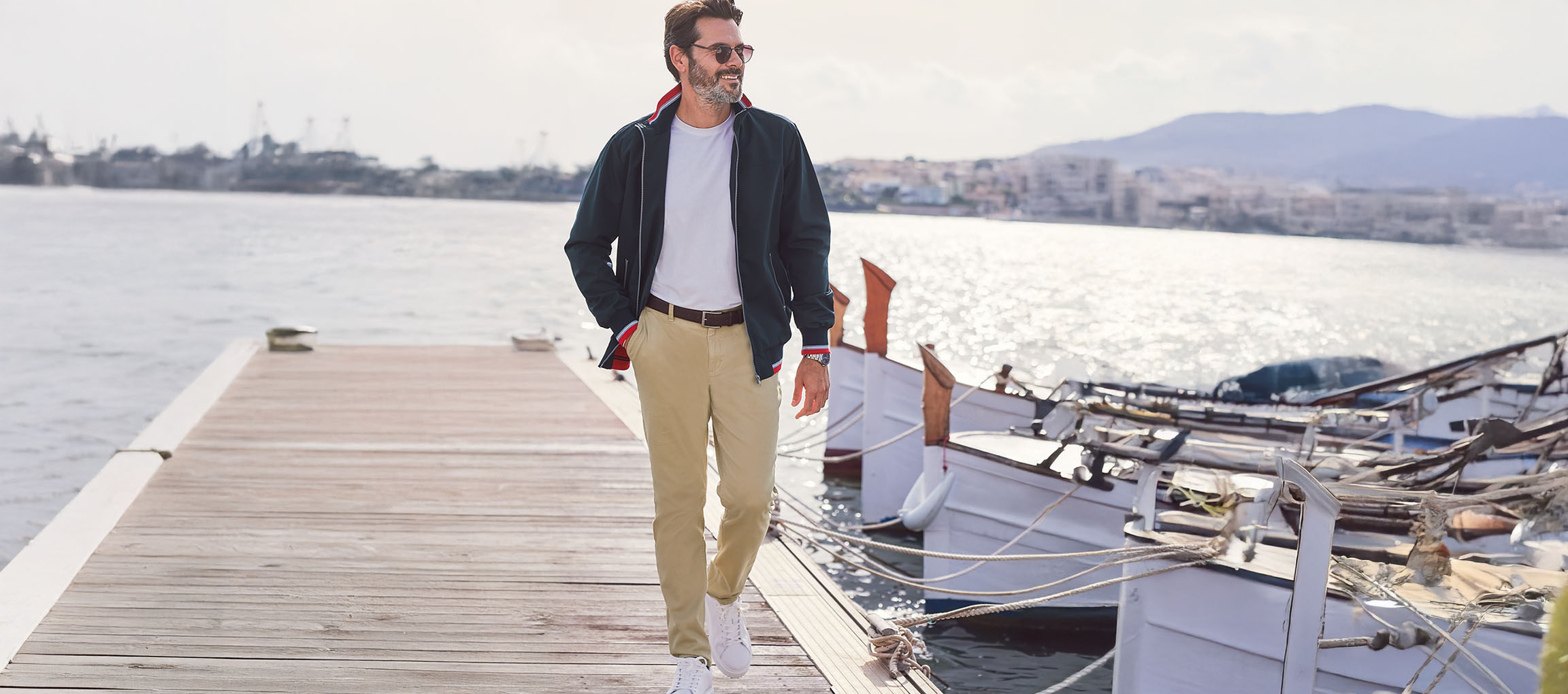 Man in a dark green bomber jacket, chino pants, and white sneakers on a dock.