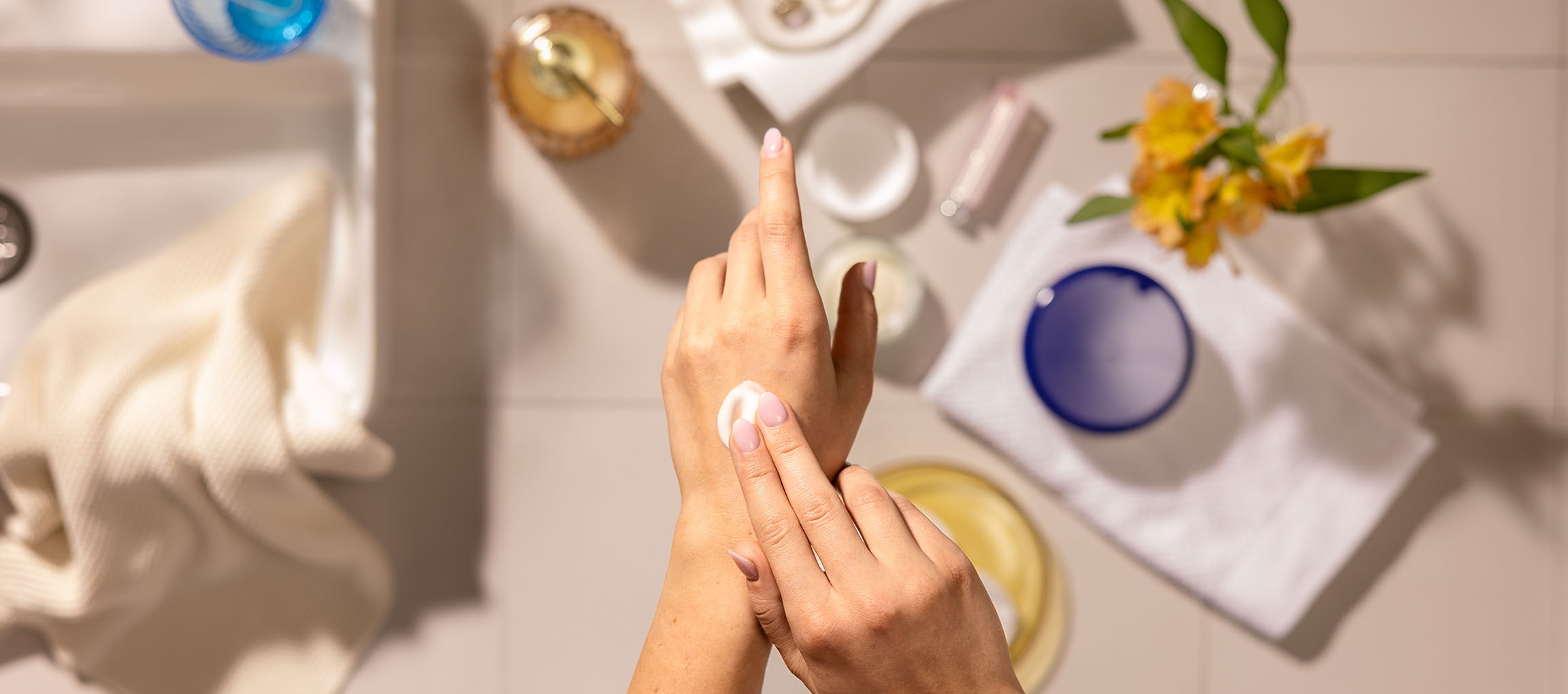 Female hands applying hand cream, surrounded by cosmetics and towels in a bathroom setting.