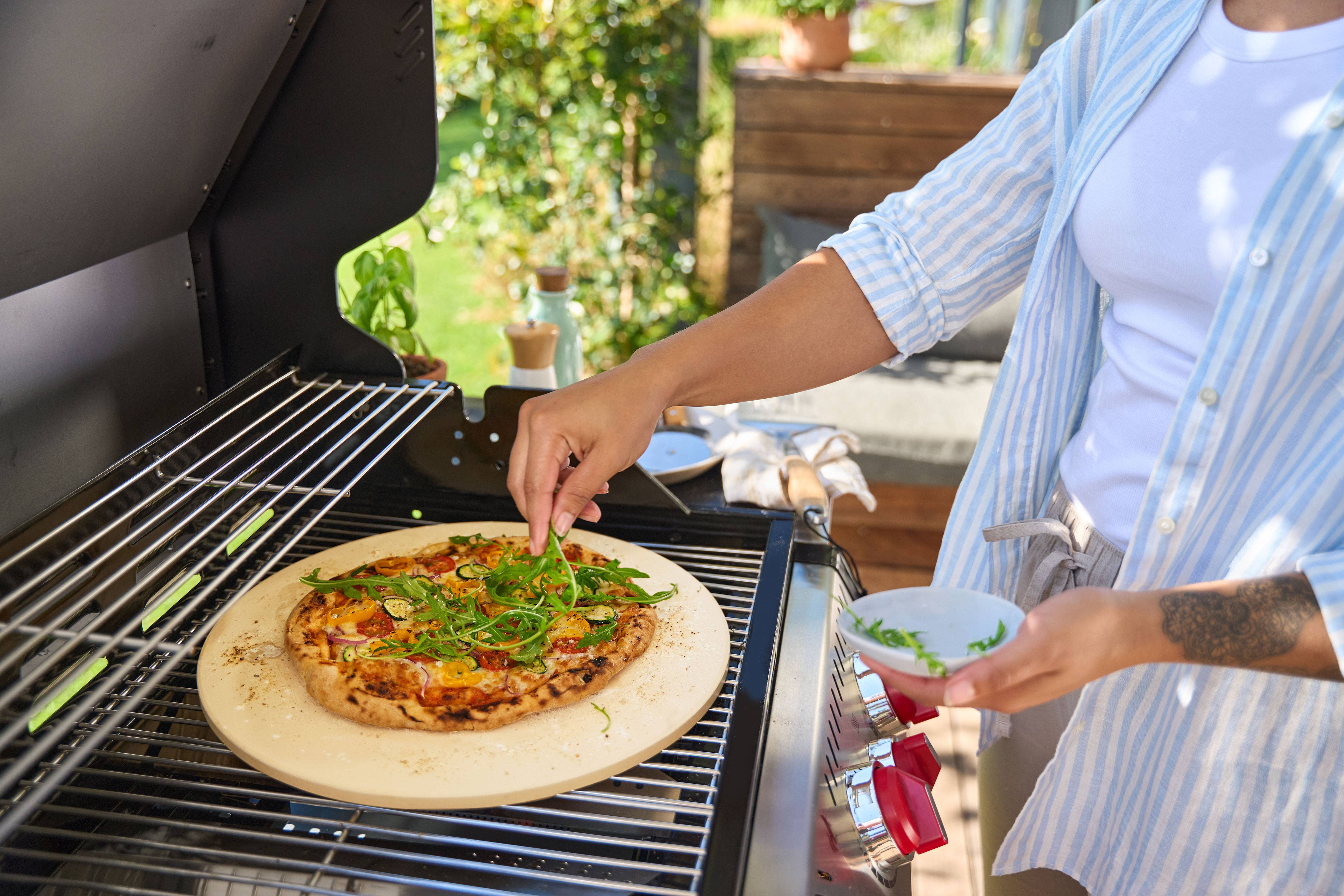 Person adding arugula to a pizza on a pizza stone in a gas grill.