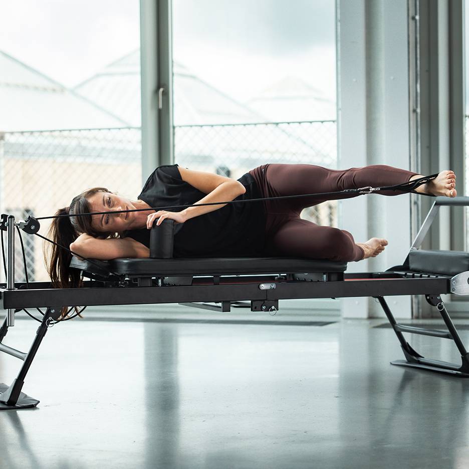 Woman doing Pilates on a reformer machine, lying on her side with leg straps.