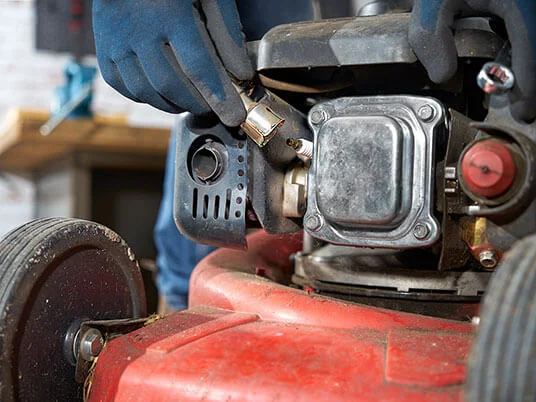 Gloved hands replacing the spark plug on a red lawnmower.