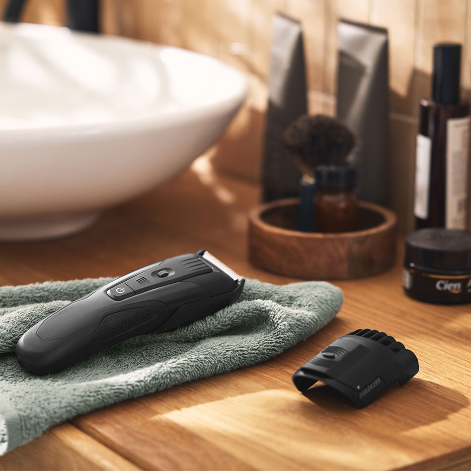 Black hair trimmer on a green towel in a bathroom with a sink and grooming products.