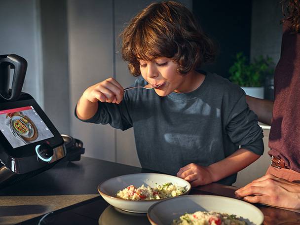 Child eating from a bowl next to a smart kitchen appliance displaying a recipe.