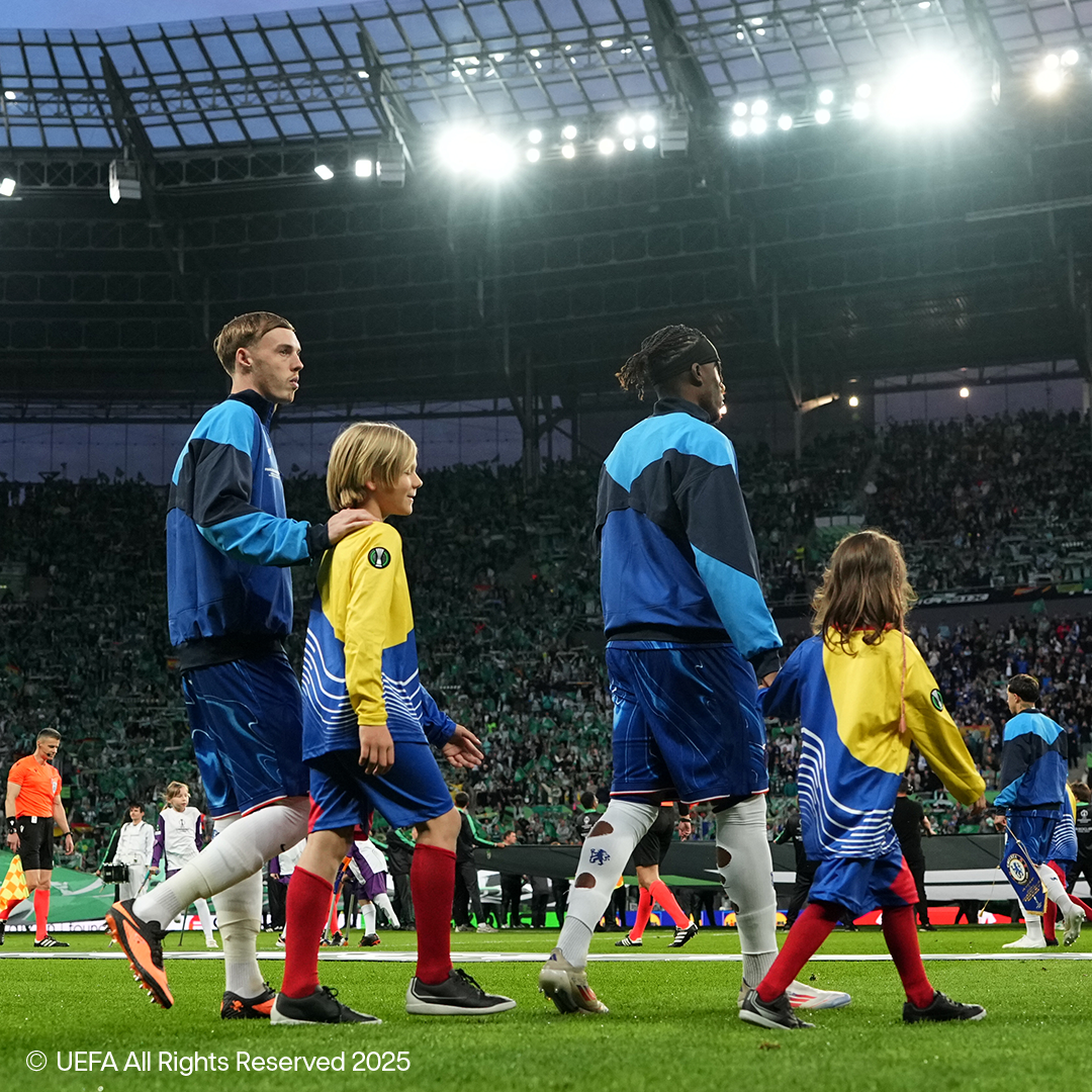 Football players and children walking on the field of a full stadium.