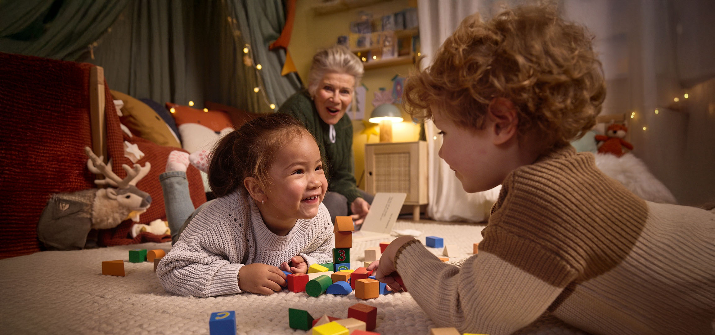 Two children playing with colorful building blocks on the floor, with an adult watching.