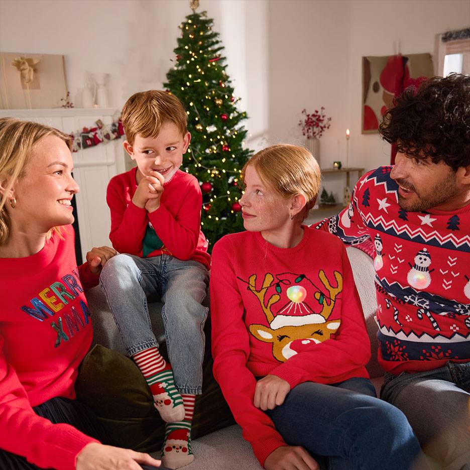 Family in red Christmas jumpers with reindeer and snowmen, sitting on a sofa in front of a Christmas tree.