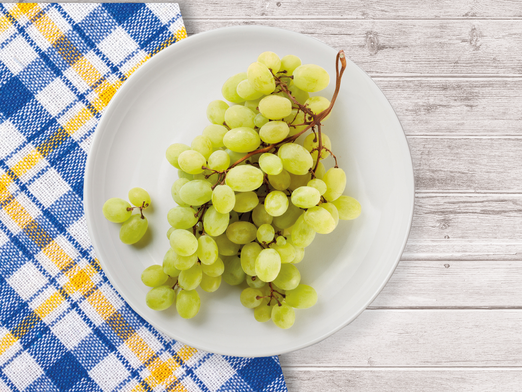 A bunch of green grapes sits on a white plate atop a blue and yellow checkered tablecloth.