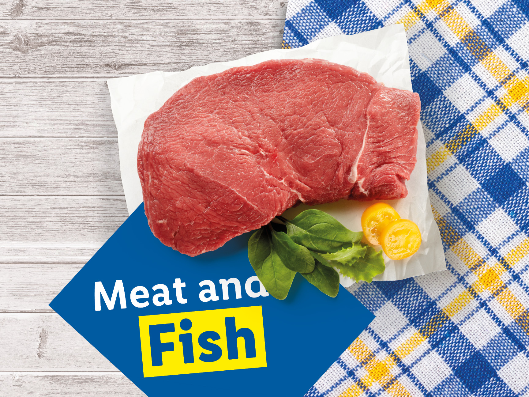 Raw beef steak with greens and tomatoes on a wooden table with a blue and yellow plaid cloth.