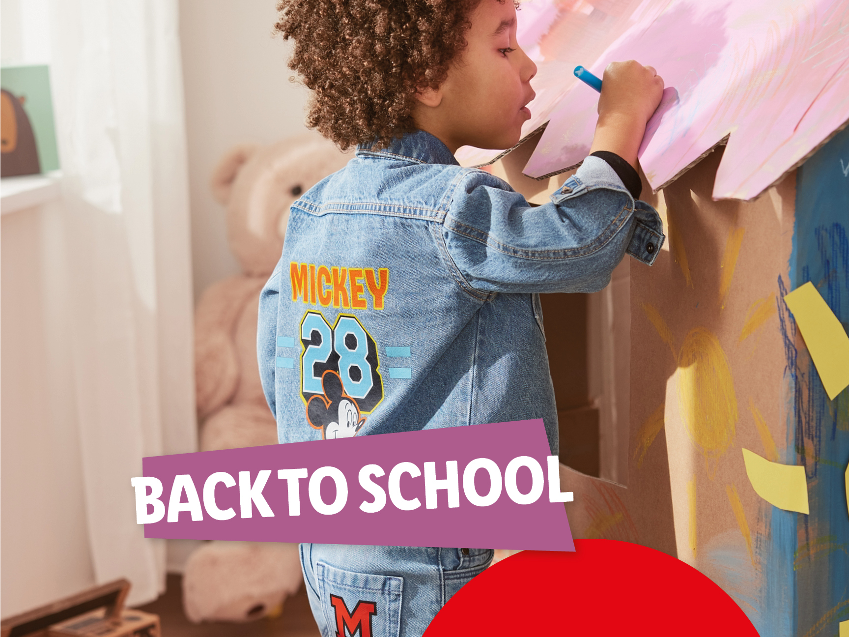 A child in a Mickey Mouse denim jacket and jeans drawing on a cardboard playhouse.