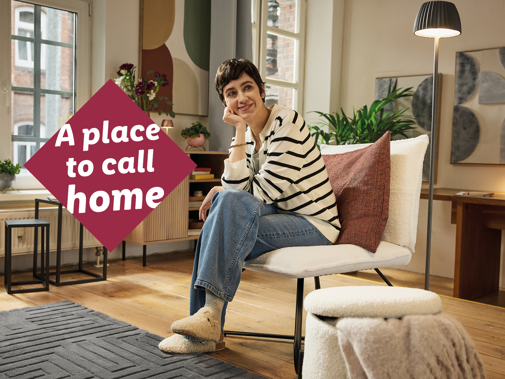 A woman in a cozy living room with a rug, chair, and floor lamp, text reads 'A place to call home'.