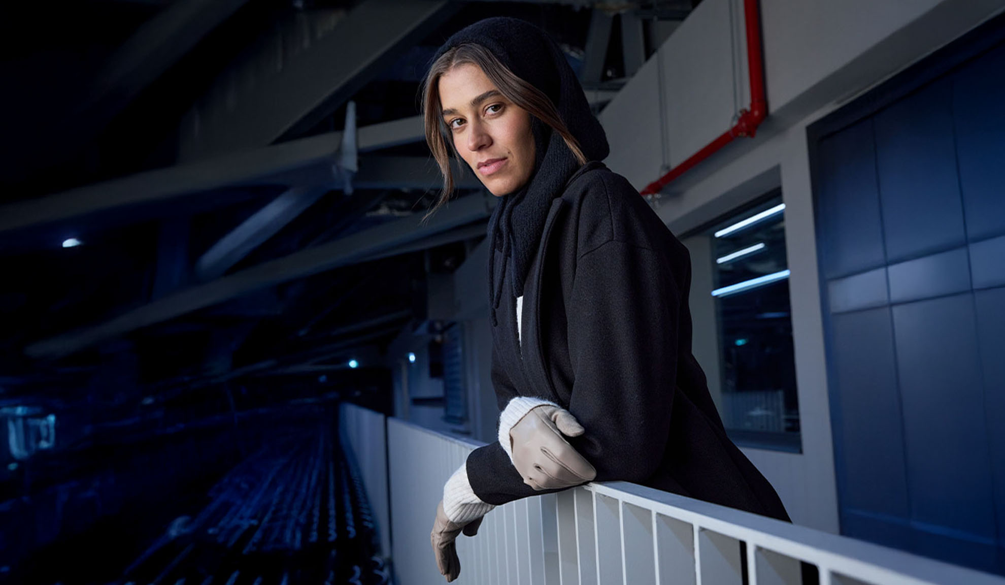 Woman in a black coat and hooded scarf, leaning on a railing in a modern setting.