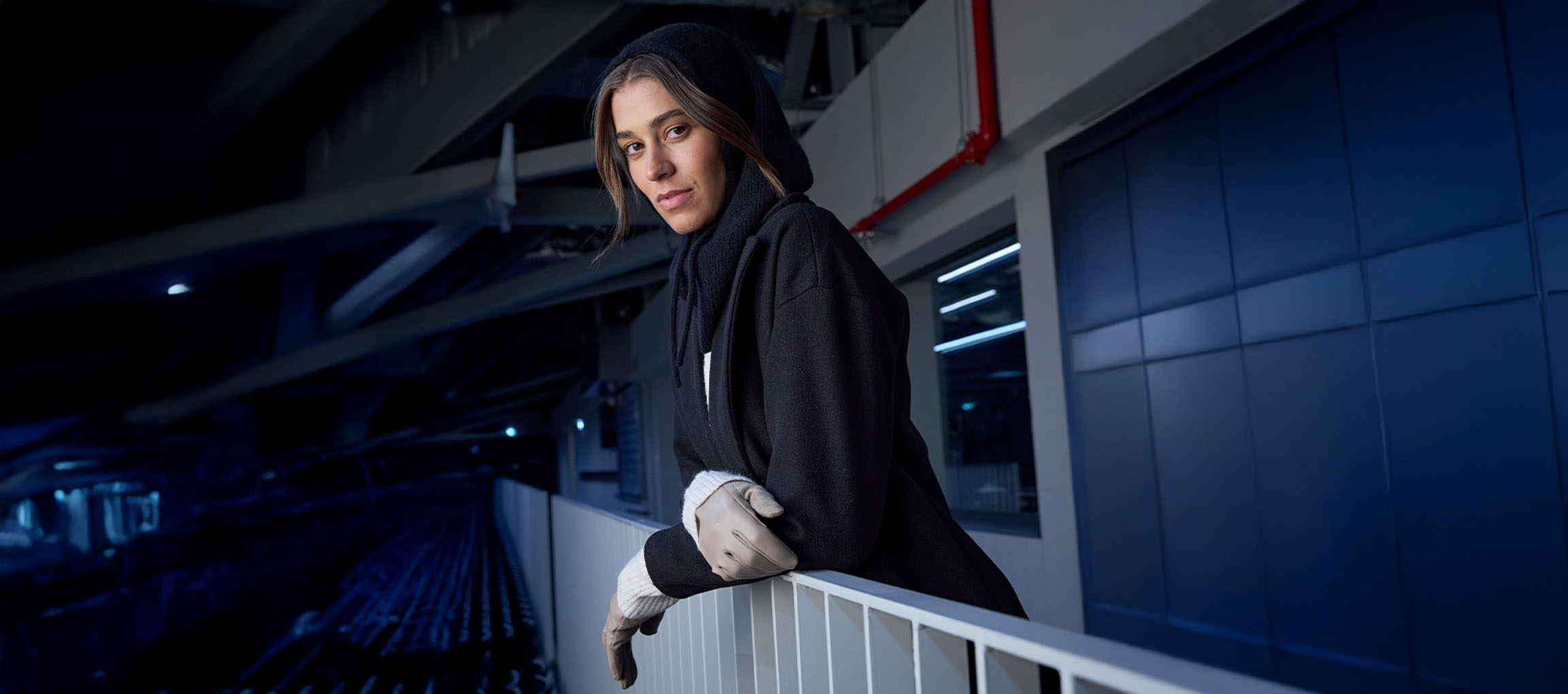 Woman in black coat and gloves leaning on a railing in an urban setting.
