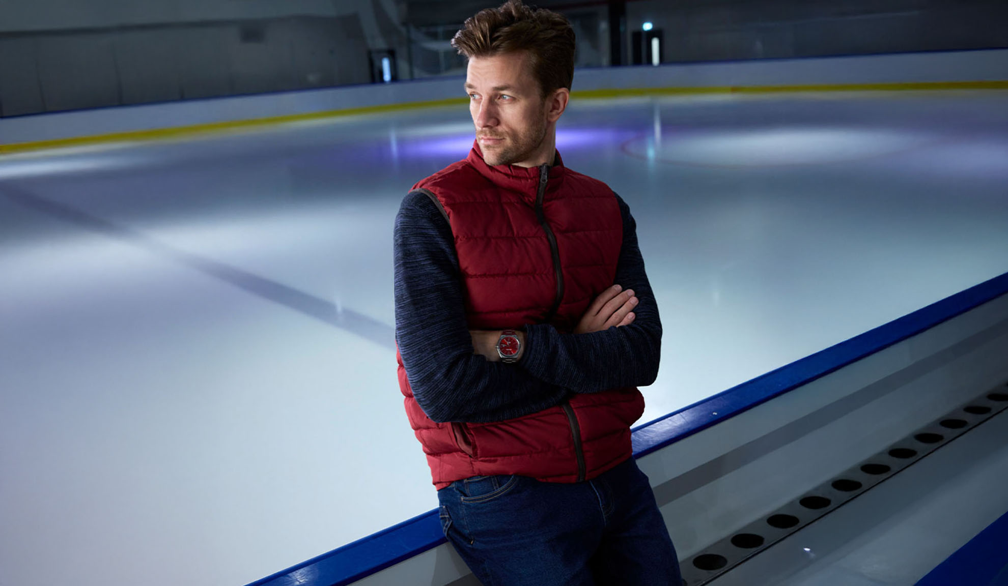 Man in a red puffer vest and blue sweater standing by an ice rink.