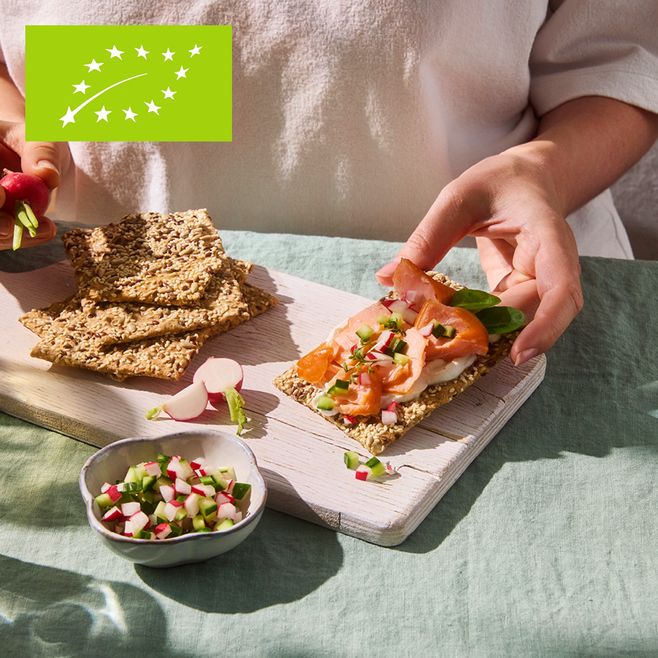 Hands preparing a crispbread with salmon, cream cheese, and diced radishes and cucumber.