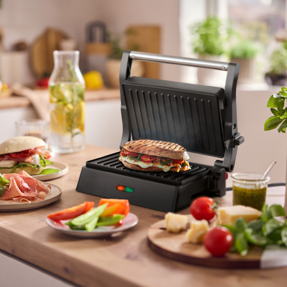Panini press with a grilled sandwich, surrounded by fresh ingredients on a kitchen counter.