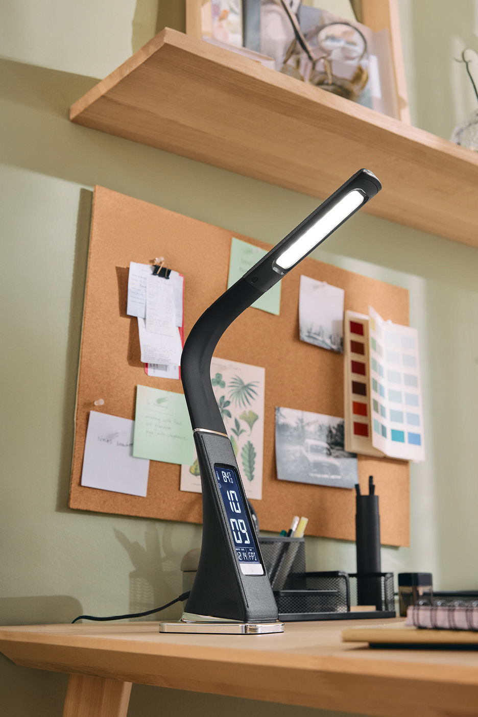 Modern LED desk lamp with digital display on a wooden desk with a cork board.
