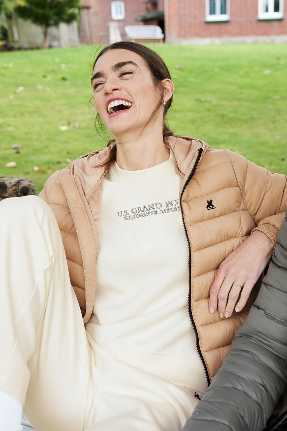 Woman in beige puffer jacket and cream sweatshirt, laughing outdoors.