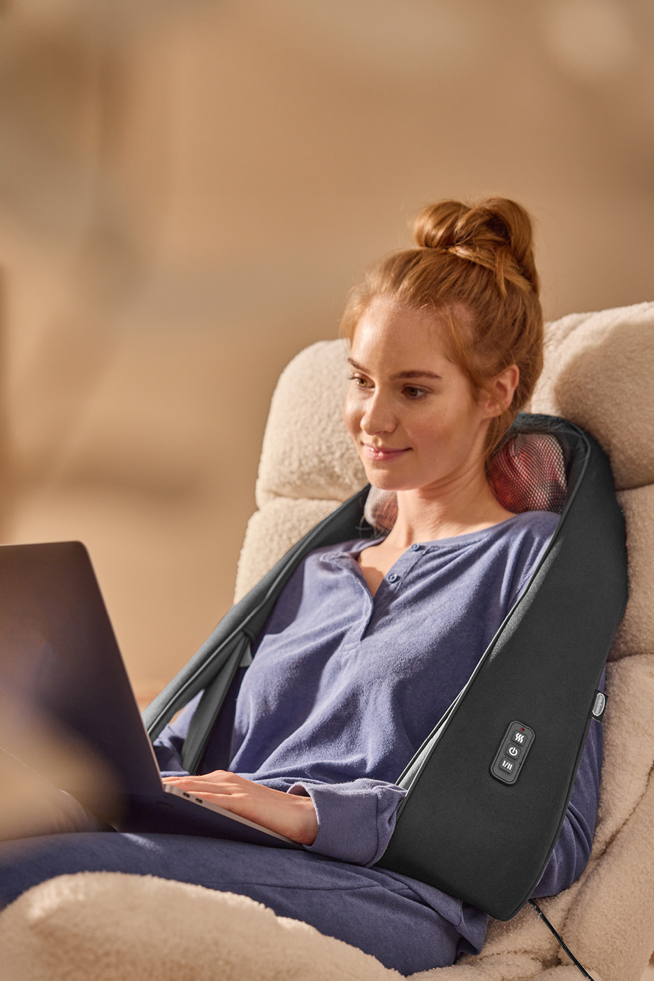 Woman using a neck and shoulder massager while working on a laptop.