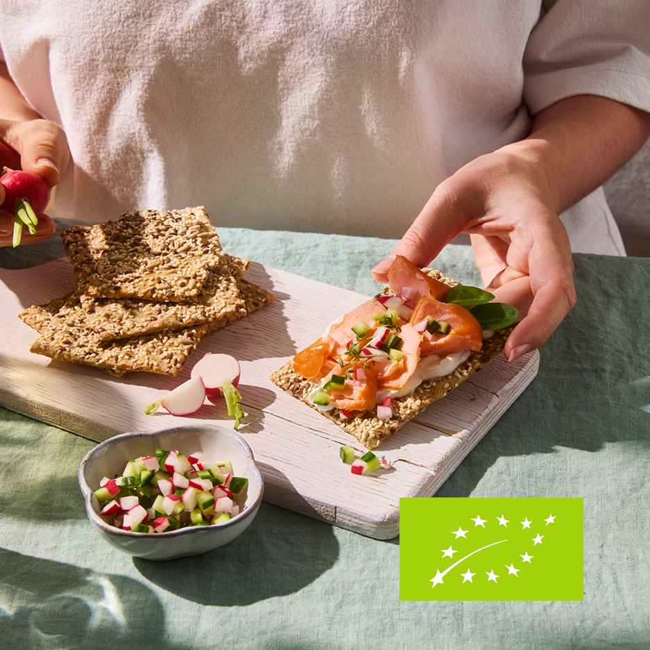 Hands preparing crispbread with salmon, cream cheese, and radish, with an organic label.