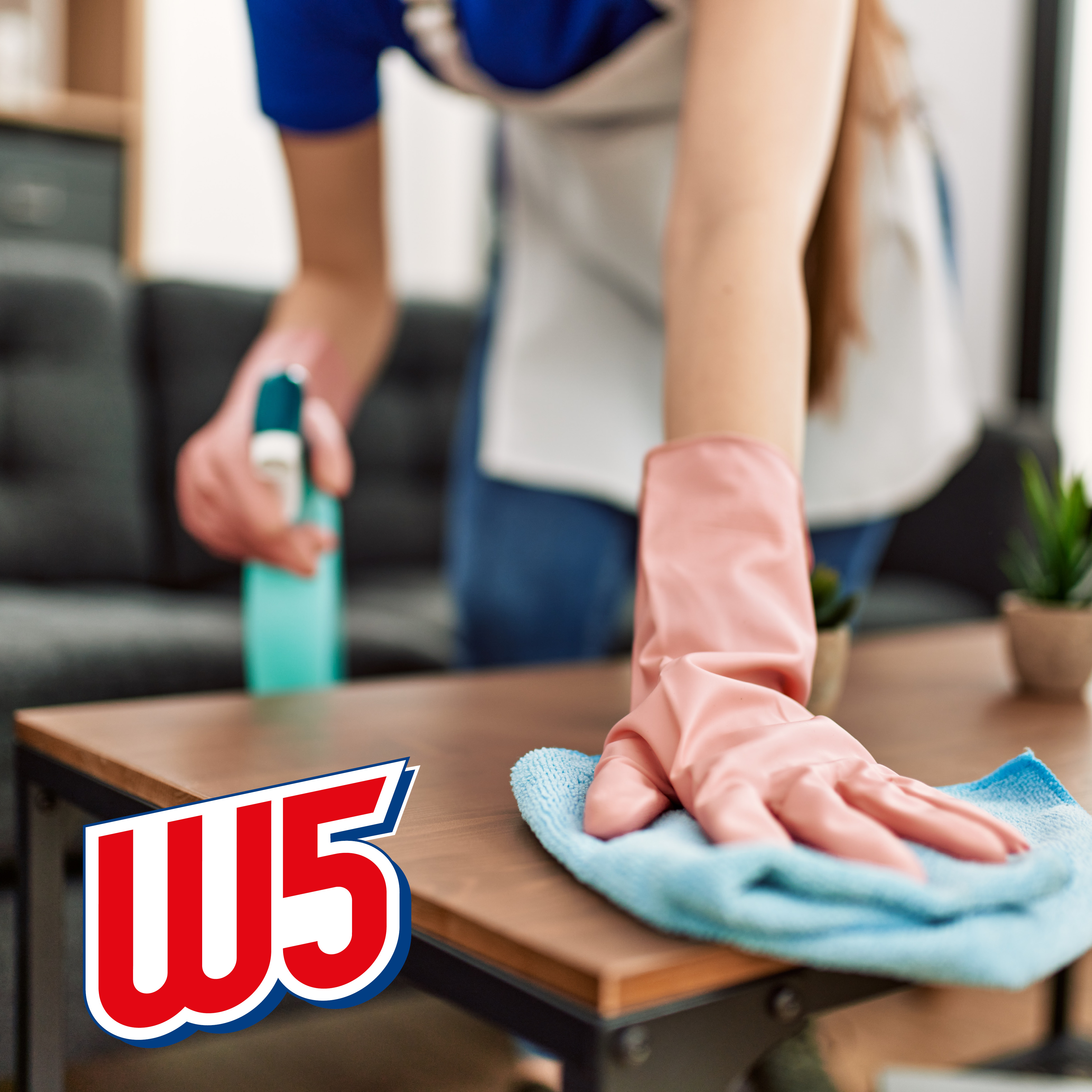 Person in gloves cleaning a wooden table with spray and cloth.