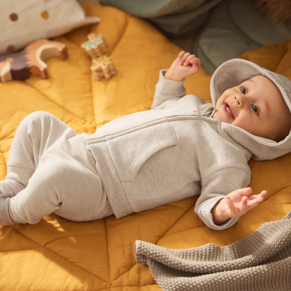 Happy baby in a grey hooded tracksuit lying on a yellow quilted blanket.