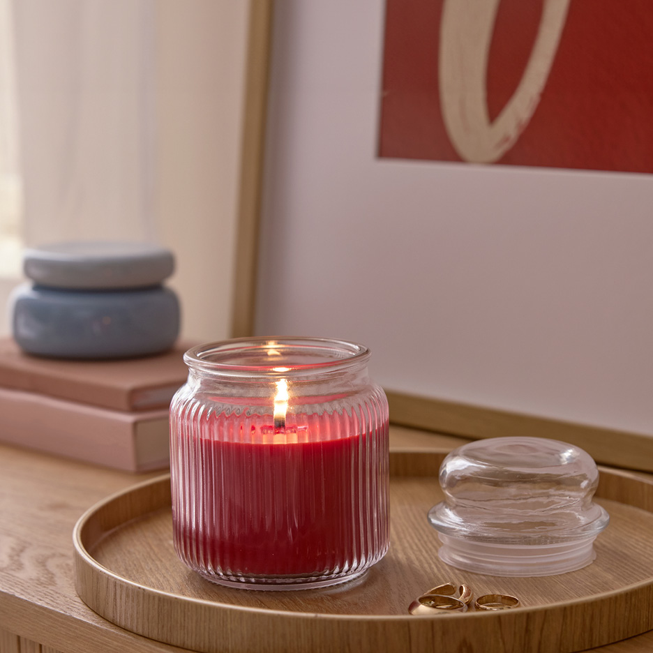 Lit red candle in a ribbed glass jar on a wooden tray with rings and a glass lid.