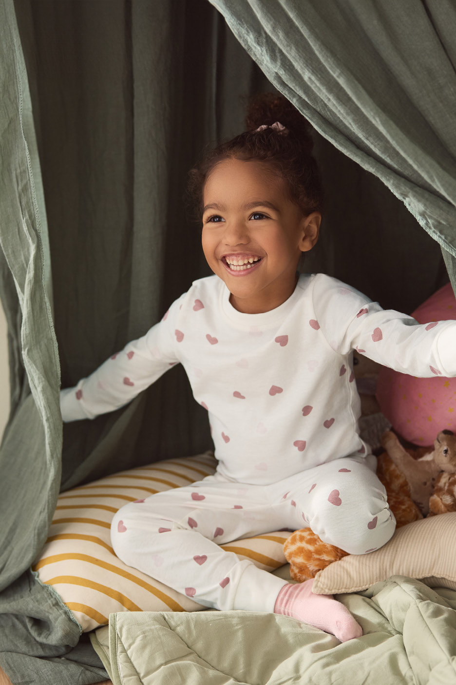 Smiling child in heart-patterned pajamas, sitting on striped cushions under a green canopy.