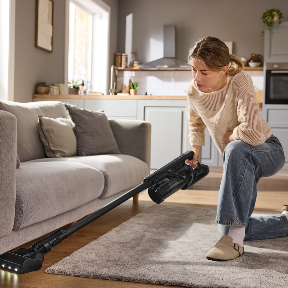 Woman vacuuming under a sofa with a cordless stick vacuum cleaner in a modern living room.