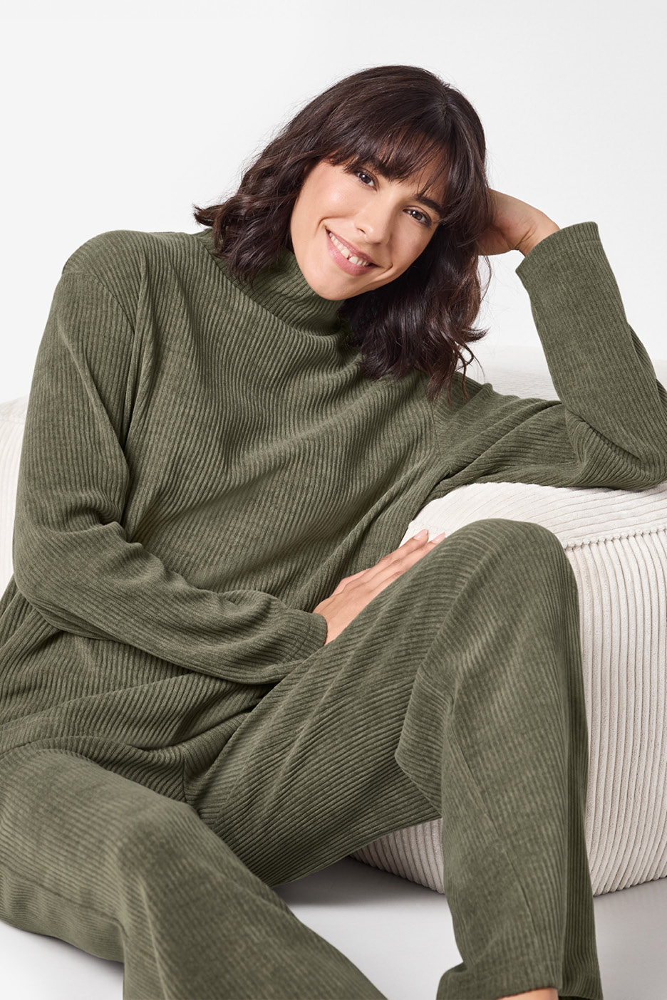 Woman in a green ribbed turtleneck and matching trousers, smiling while sitting on a white ottoman.