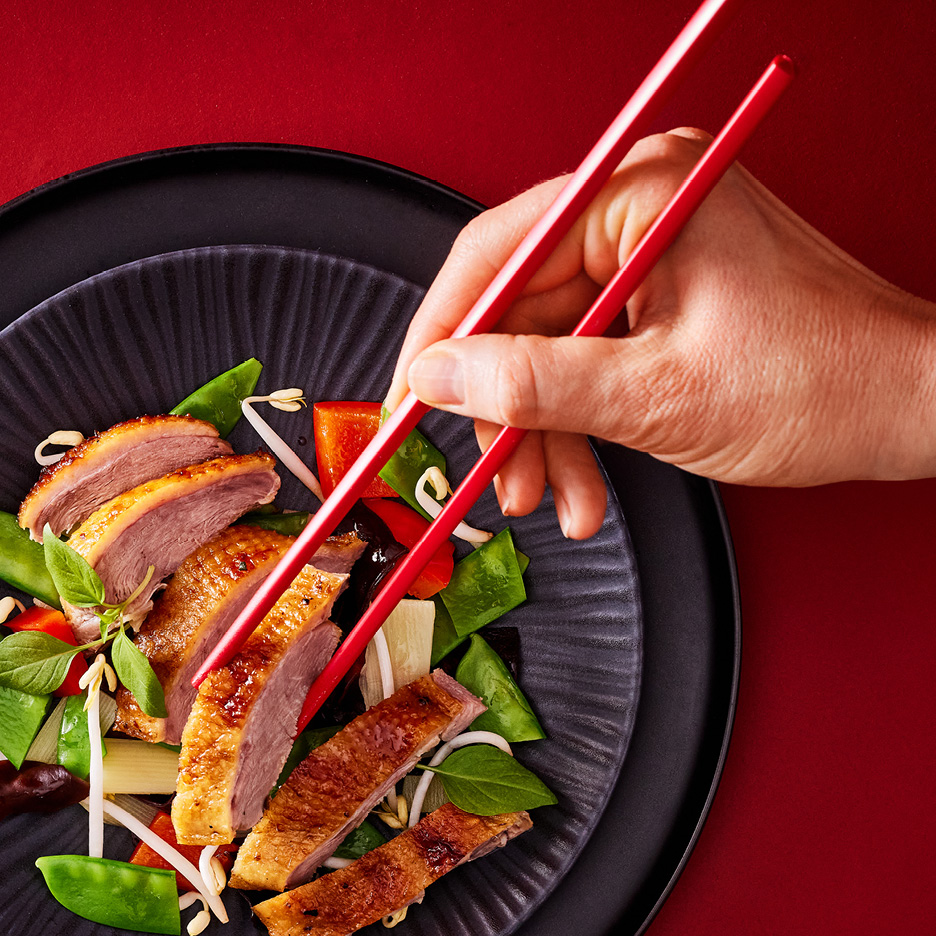 A hand holding red chopsticks over a black plate of sliced duck breast with vegetables.