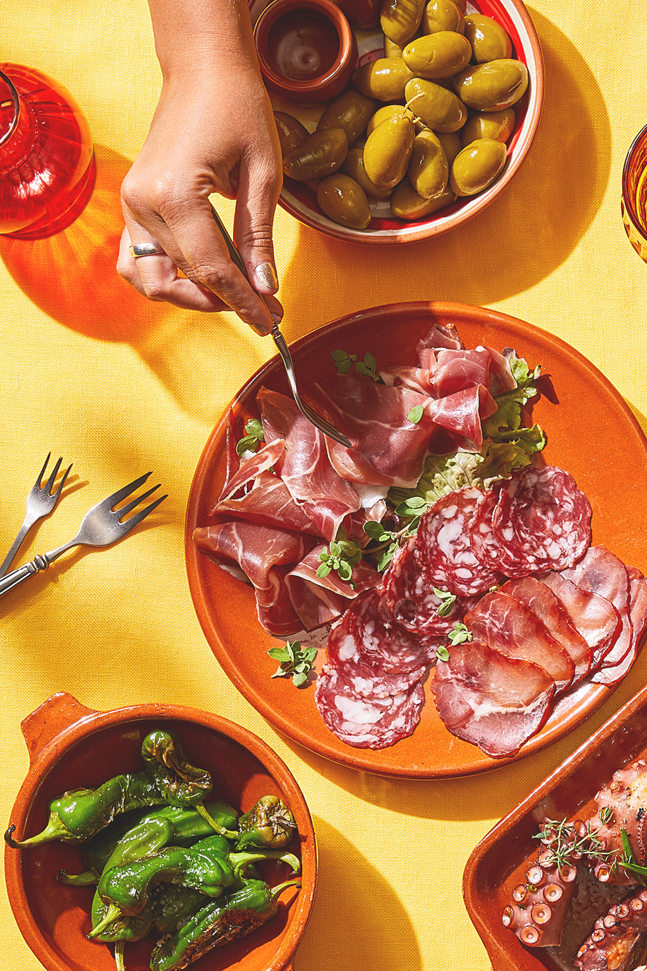 A hand forks cured meats, with olives, padrón peppers, and octopus on a yellow tablecloth.