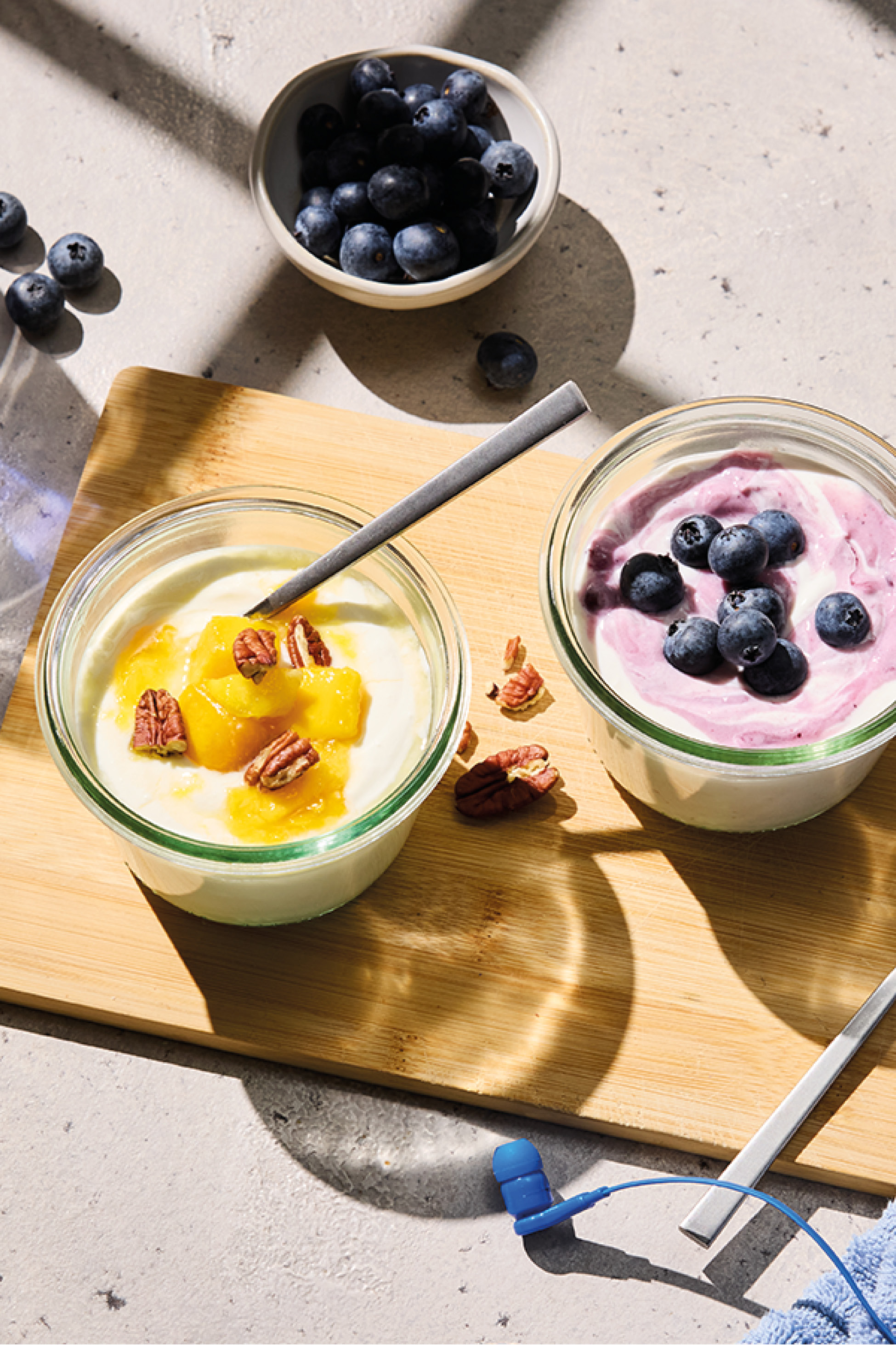 Two bowls of yogurt, one with mango and pecans, the other with blueberries, on a wooden board.