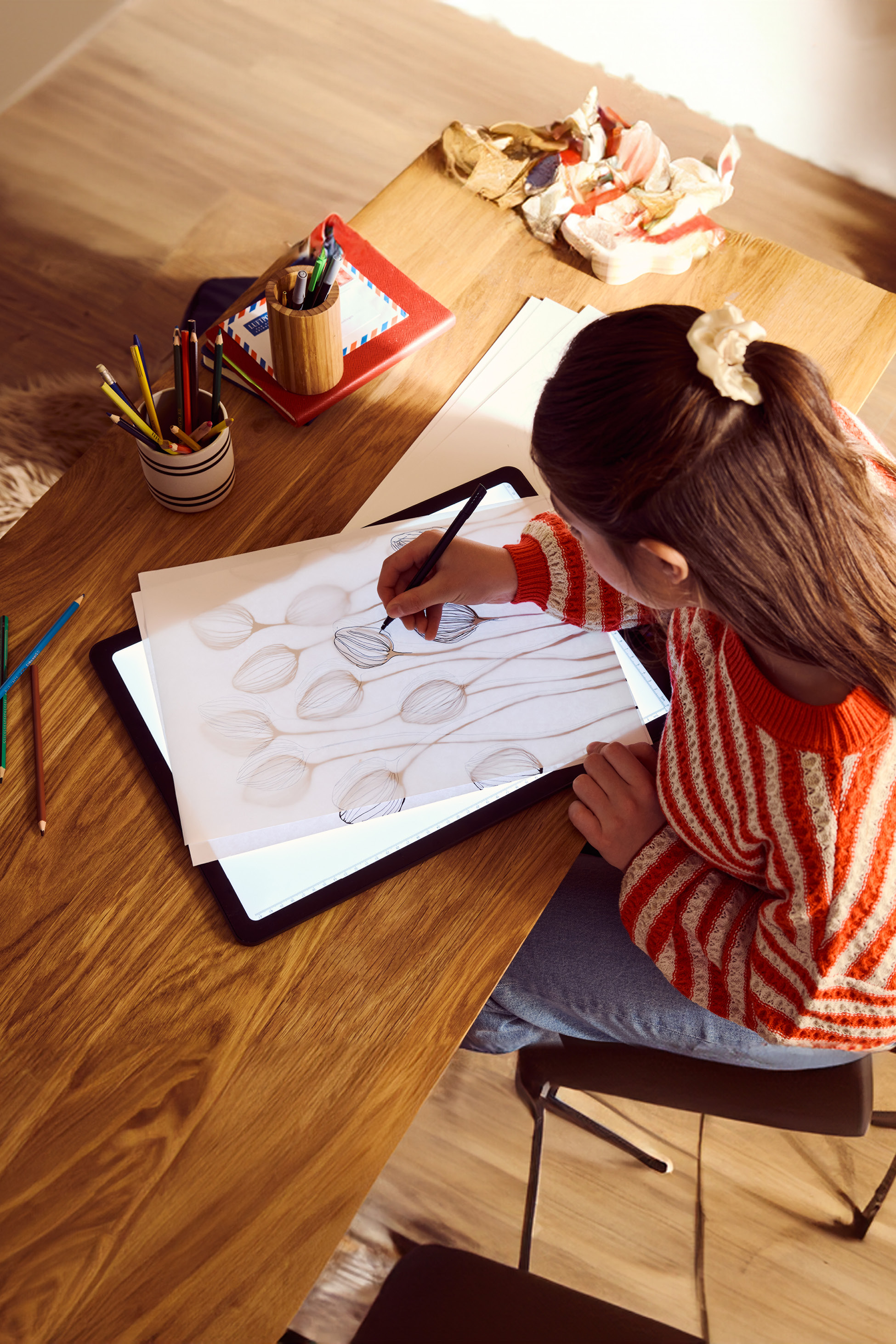 A person drawing on a light pad with a pen, surrounded by art supplies on a wooden table.