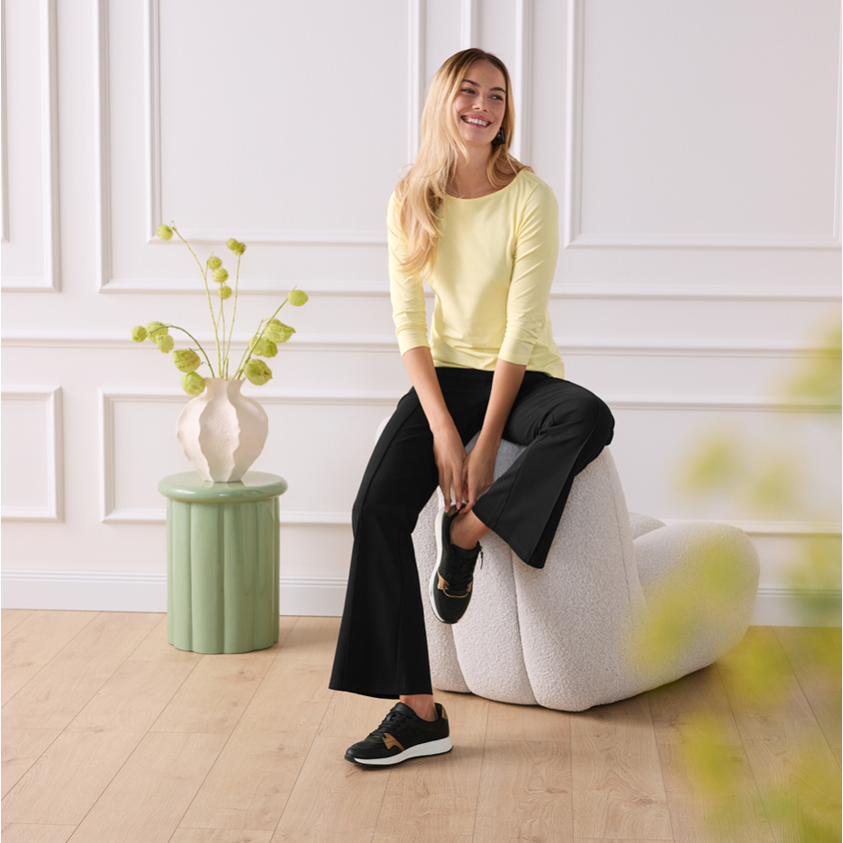 Woman in yellow long-sleeve top and black trousers sitting on a white chair.